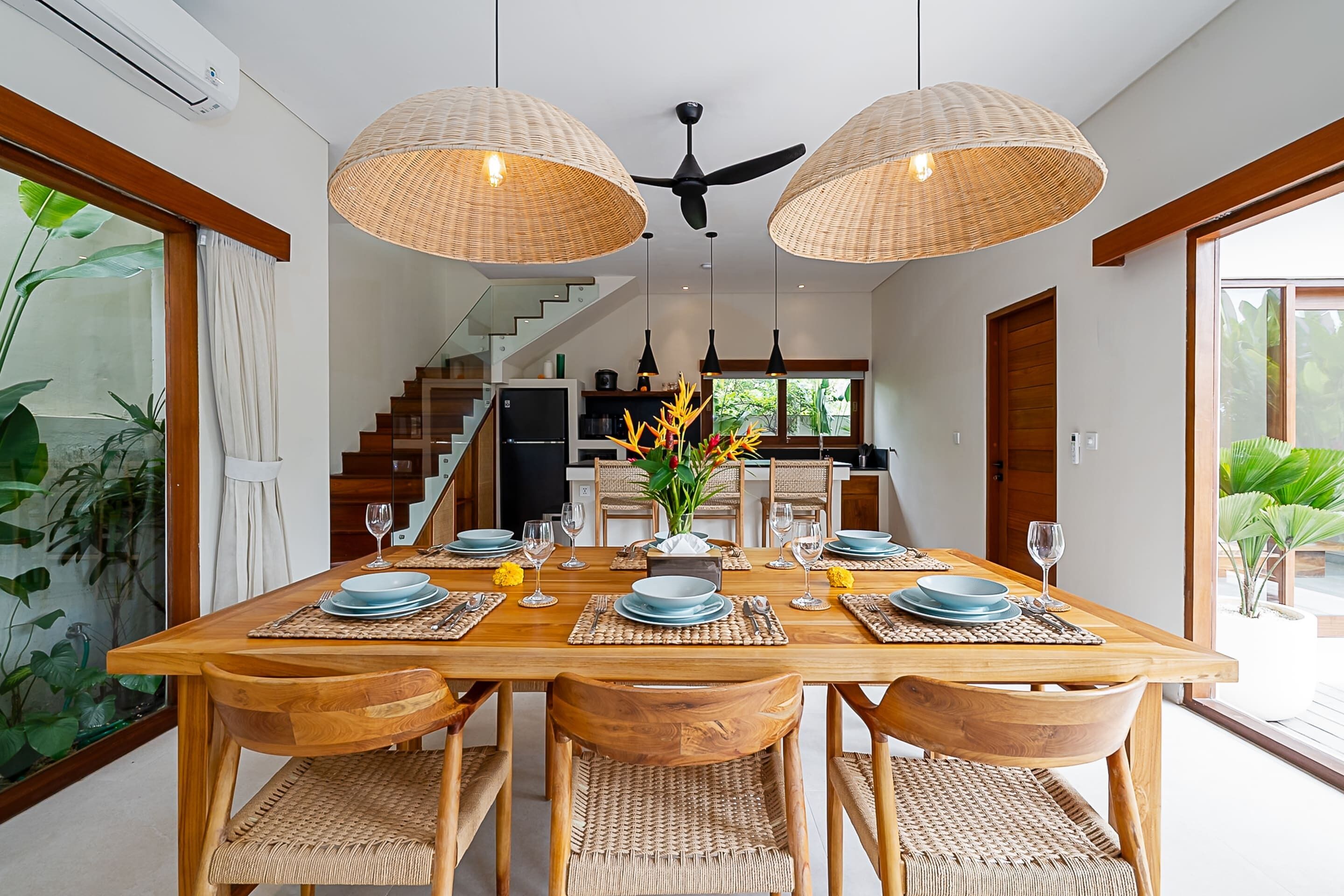 A dining area with a wooden table set for six, surrounded by wicker chairs, with a ceiling fan and woven light fixtures above.