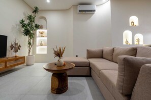 Ein modernes Wohnzimmer mit einem beigefarbenen Sofa, einem Holztisch, einem Fernseher auf einem Holzständer, einer Topfpflanze und einer Wand-Klimaanlage.