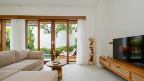A modern living room with a sofa, a coffee table, a TV, and a sliding glass door leading to a pool area. - Villa Yen by Elysian (Kerobokan)
