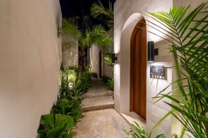 A narrow pathway flanked by lush greenery and a white building with a wooden door. - Villa Segia by Elysian (Kerobokan)