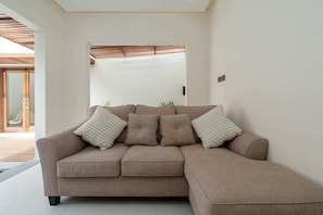 A modern living room with a beige sectional sofa, a wooden ceiling, and a glass door leading to an outdoor area. - Villa Segia by Elysian (Kerobokan)
