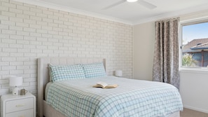 A bedroom with a white brick wall, a bed with checkered bedding, a nightstand with a lamp, and a window with curtains. - Barefoot Beach House - Lifestyle Yamba (YAMBA)