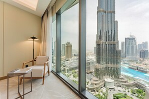 A modern room with a large window, a chair, a small table, and a view of a cityscape with a prominent skyscraper. - PVH - Address Opera 3BR (Dubai)
