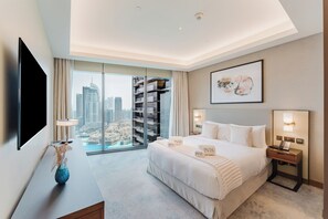 A hotel room with a large bed, a flat-screen TV, a desk with a lamp, a vase, a city view through a glass door, and a framed artwork on the wall. - PVH - Address Opera 3BR (Dubai)