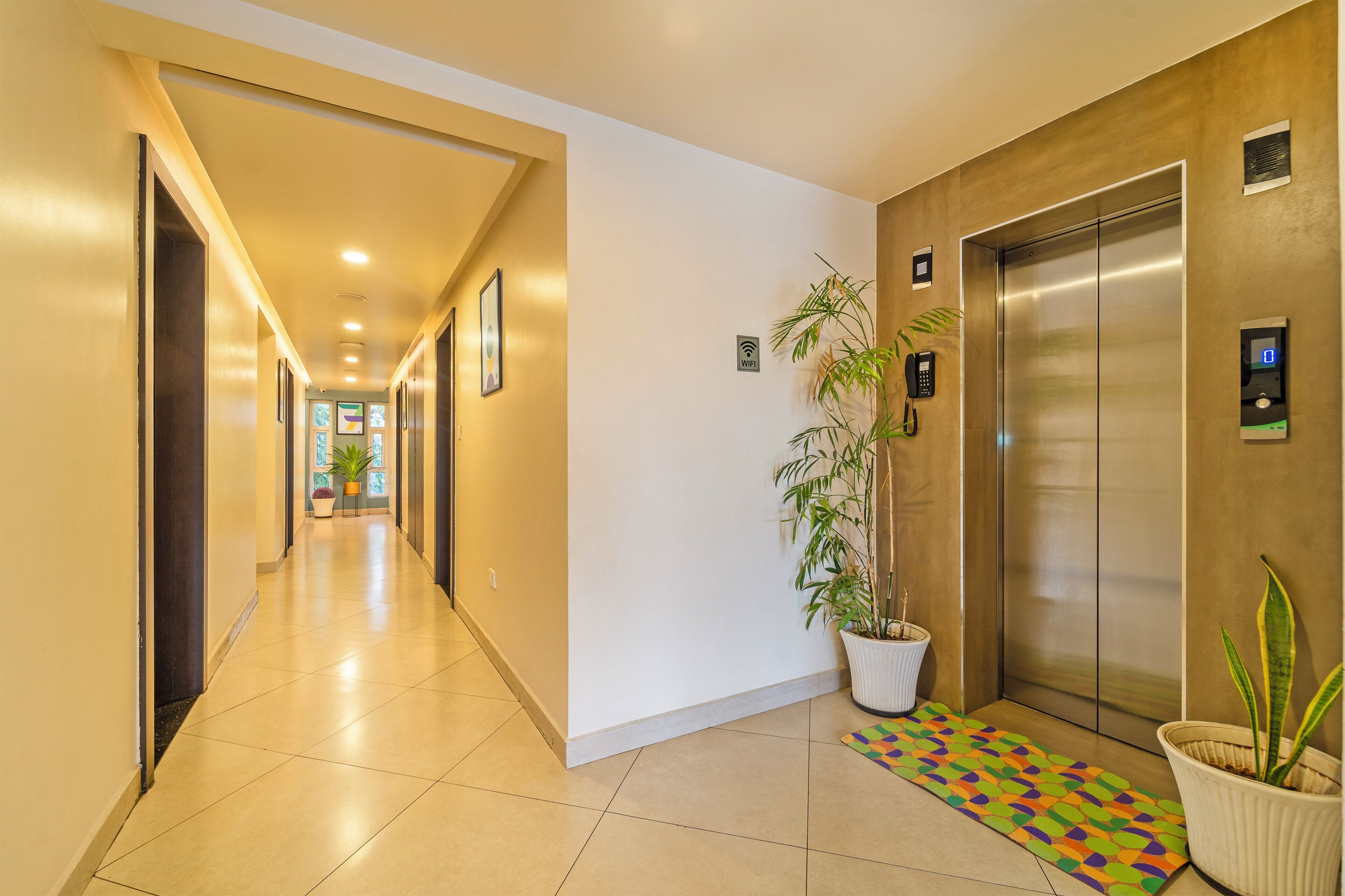 Un passadís d'hotel amb sòl de rajoles, una planta en test, un ascensor i una catifa colorida.