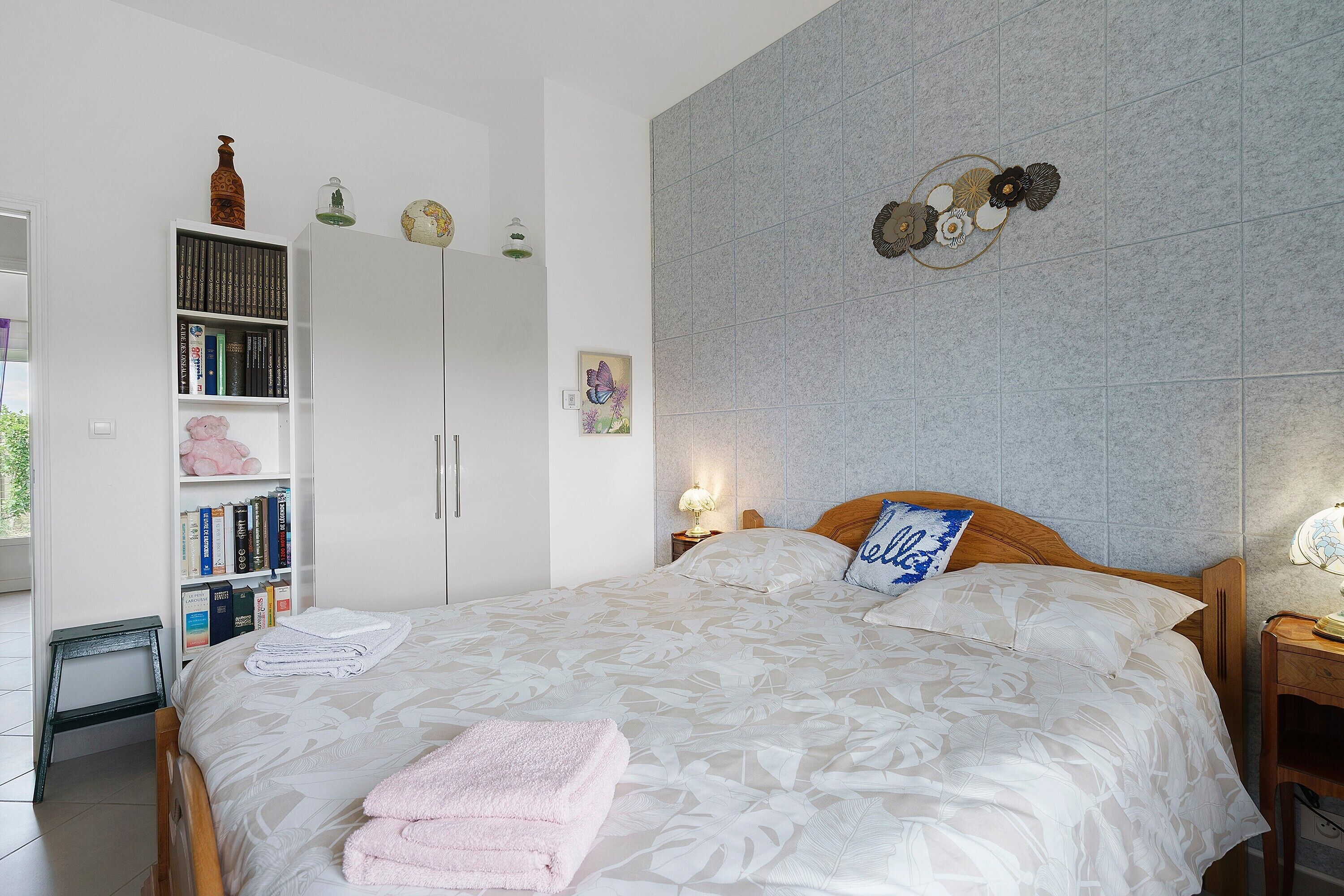 Ein ordentlich bezogenes Bett mit weißer blumiger Tagesdecke, ein rosafarbenes Handtuch und ein blaues Kissen mit Blumenmuster. An der Wand hängt ein dekorativer Teller, daneben ein Bücherregal mit Büchern und Dekorationsobjekten sowie ein Nachttisch aus Holz mit einer Lampe.