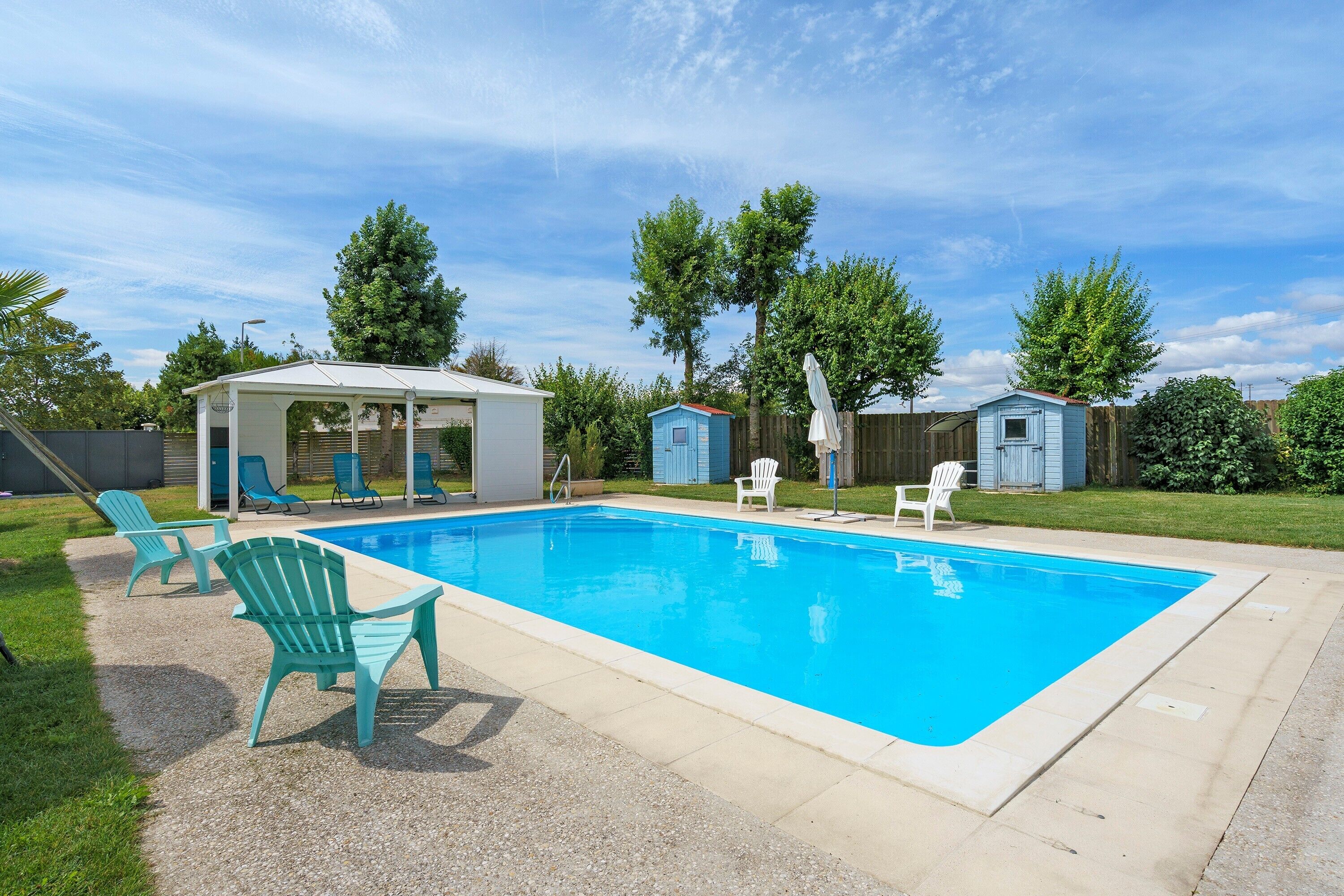 Une piscine rectangulaire dotée d’un patio couvert, de chaises d’extérieur et d’un petit abri de jardin.