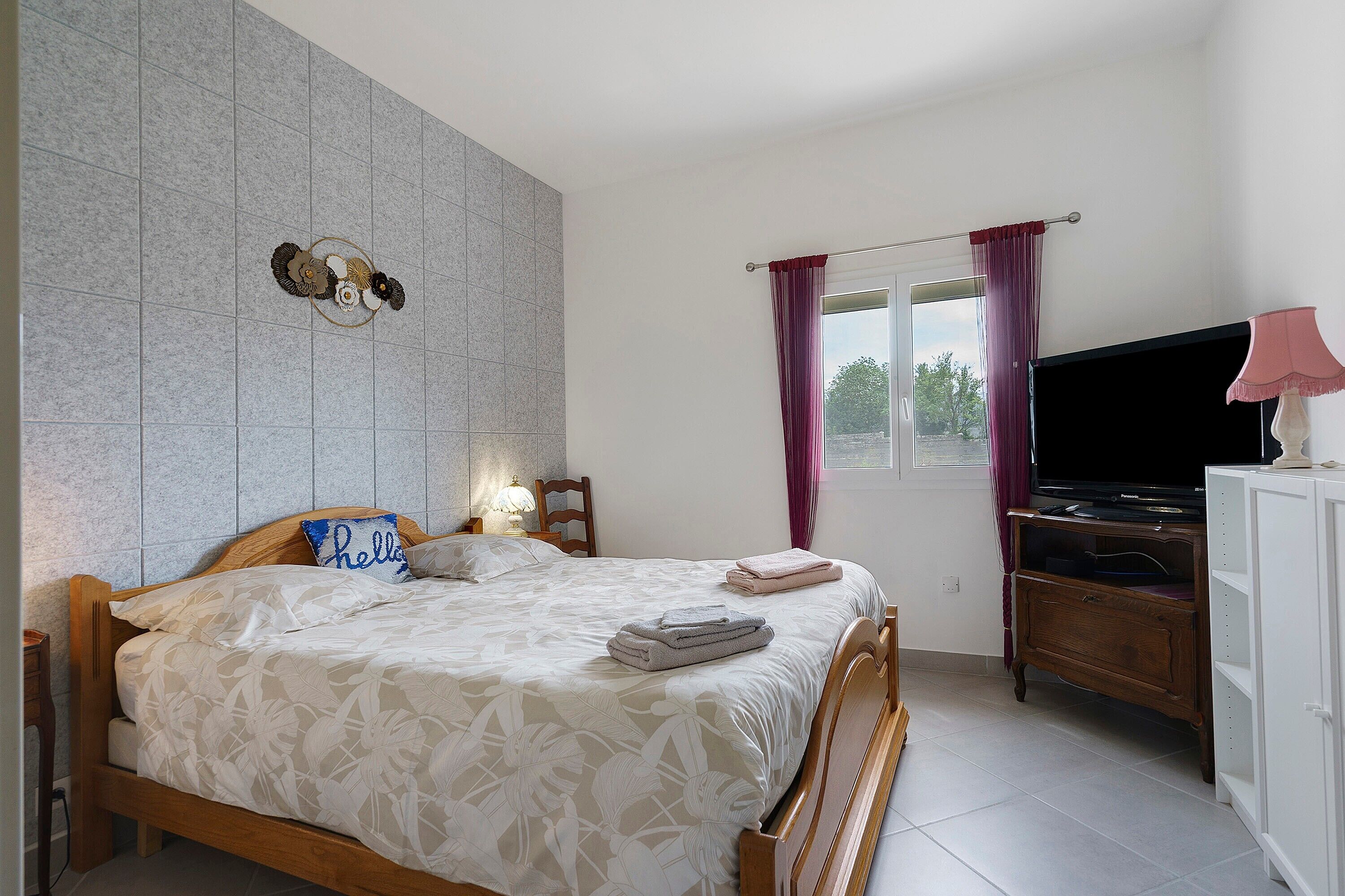 Een slaapkamer met een bed, een televisie, een dressoir en een raam met gordijnen.