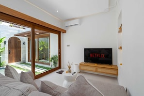 A modern living room with a TV, sofa, and a glass door leading to a garden. - Villa Numa by Elysian (Kerobokan)