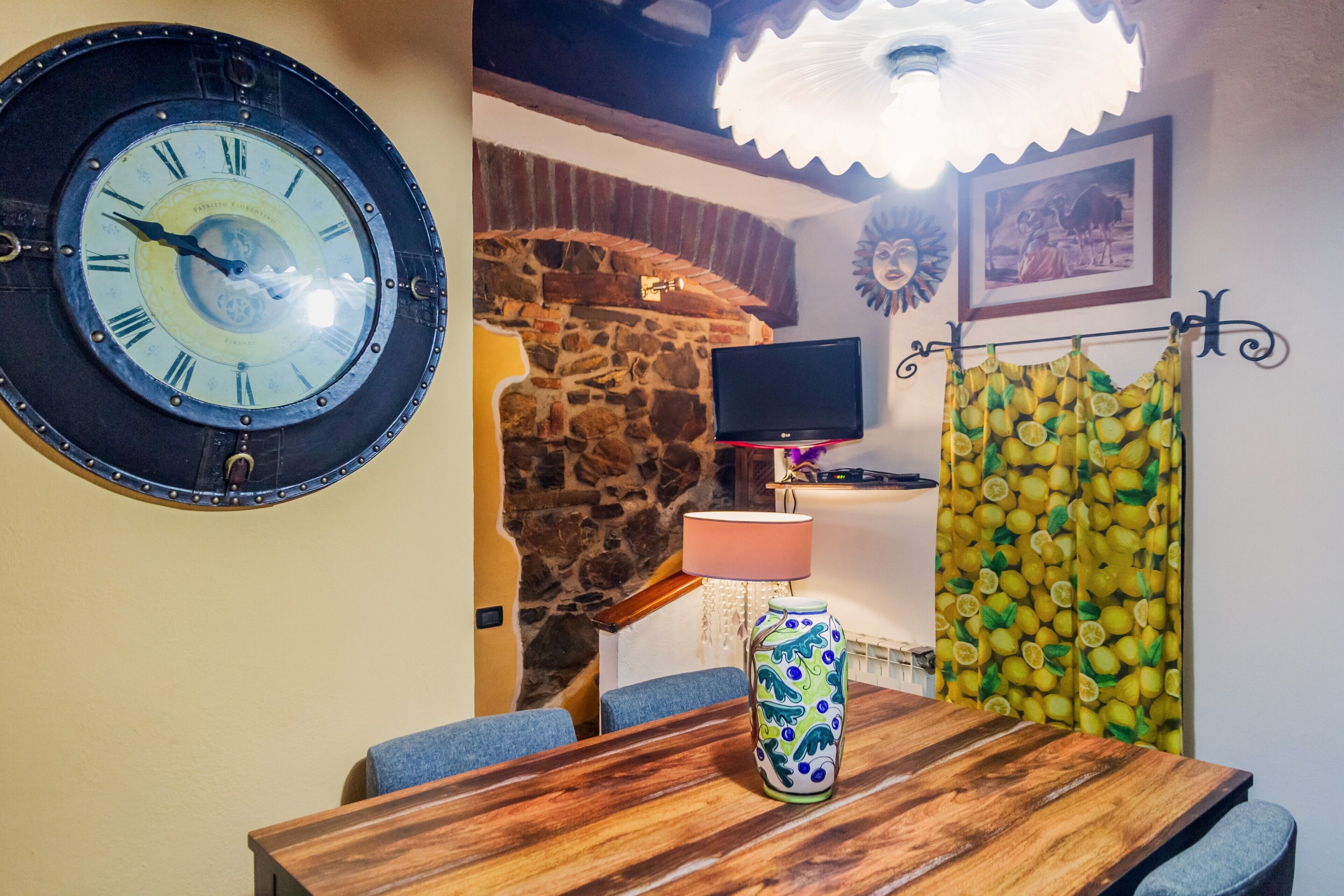 Une pièce avec un mur en pierre, une table en bois, une horloge, un téléviseur et un rideau à motifs.