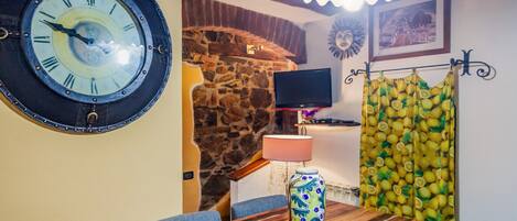 Une pièce avec un mur en pierre, une table en bois, une horloge, un téléviseur et un rideau à motifs.