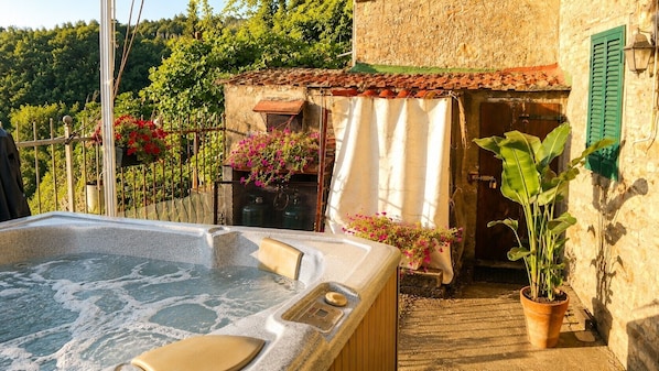 Un bain à remous avec un revêtement extérieur en pierre, une terrasse en bois et un bâtiment rustique doté d’un toit en tuiles et de volets verts.
