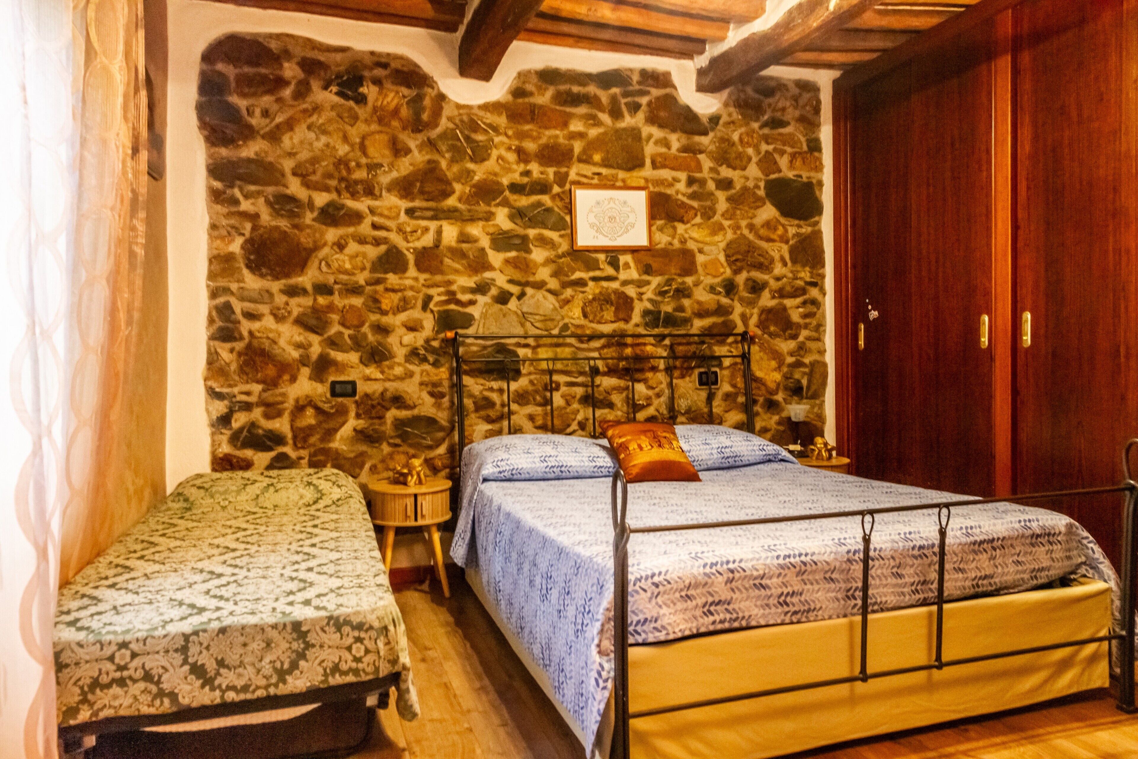 A bedroom with a stone wall, a wooden door, a bed with a metal frame, and a small bedside table.
