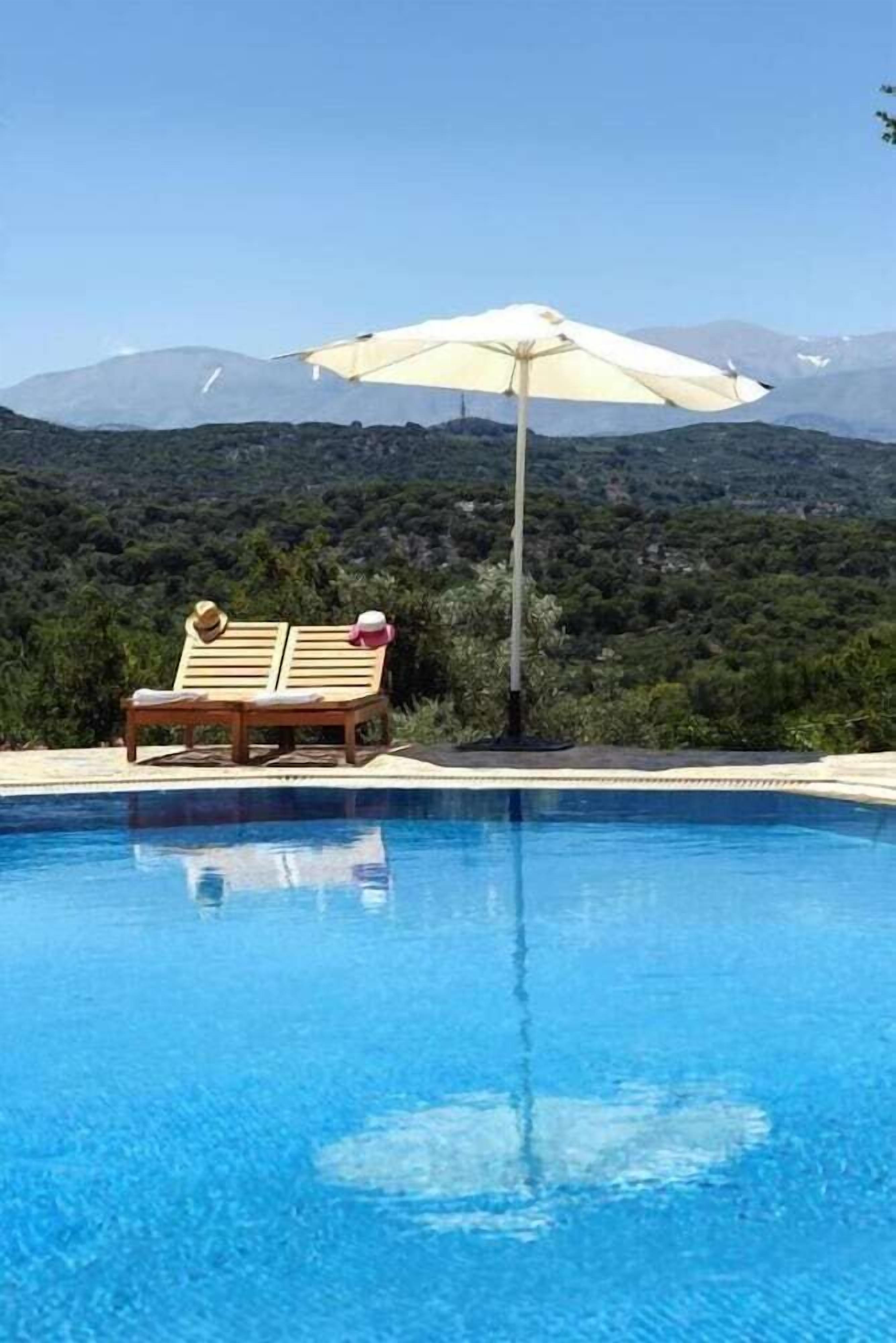 Une piscine avec un fauteuil de relaxation en bois et un parasol, entourée de verdure et de montagnes.