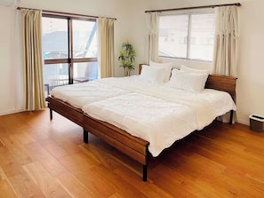 Ein Schlafzimmer mit einem hölzernen Bett, großen Fenstern mit Vorhängen, einer Topfpflanze und Blick auf Gebäude.