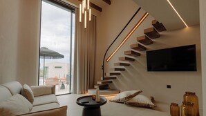 A modern living room with a sofa, a coffee table, a TV mounted on the wall, a staircase with wooden steps, and a large window. - Standard Villa - Kyklio Pavilions Nafplio (Nafplio)