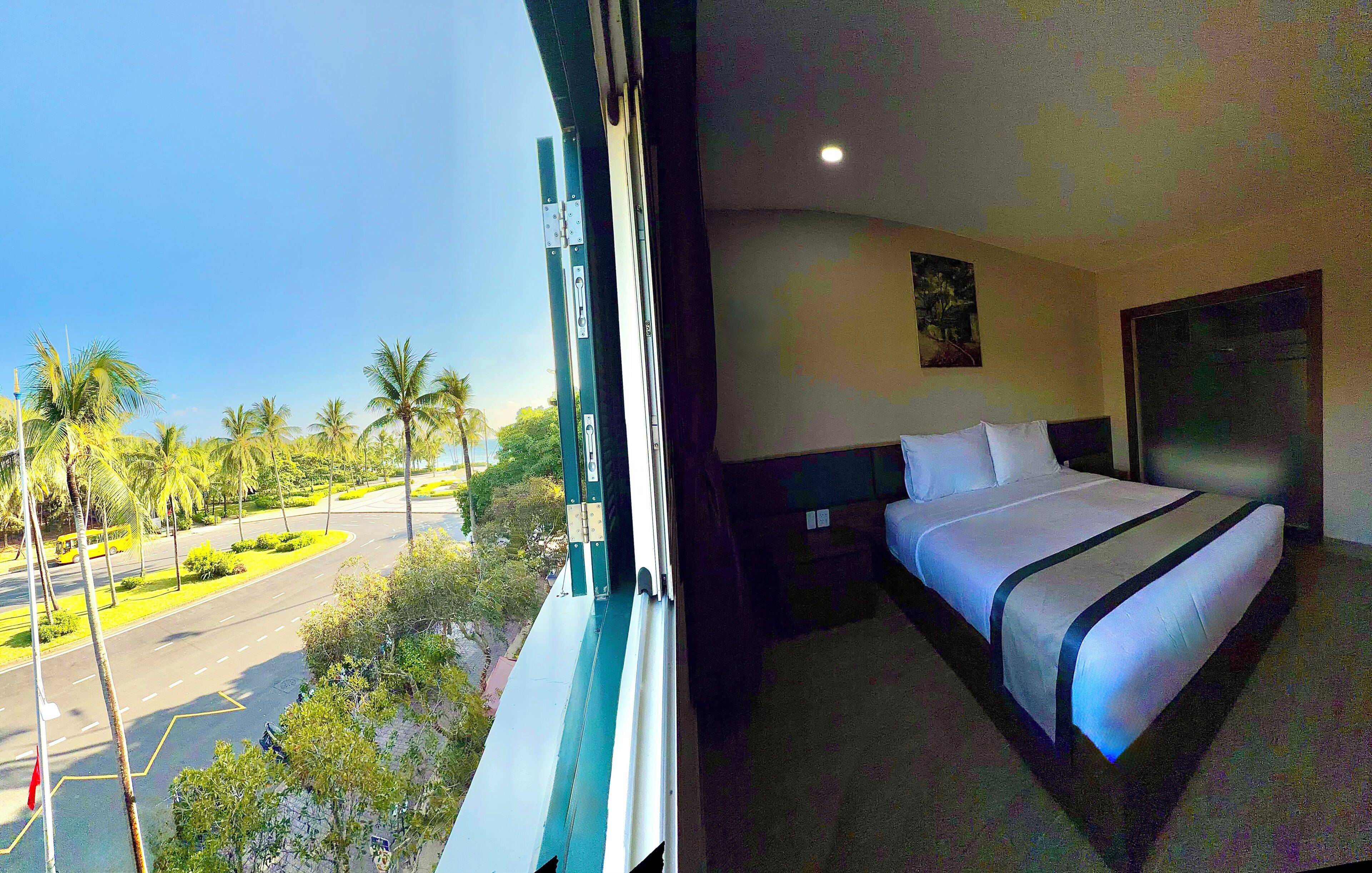 A hotel room with a large bed, a nightstand, a window with a view of palm trees and a road.