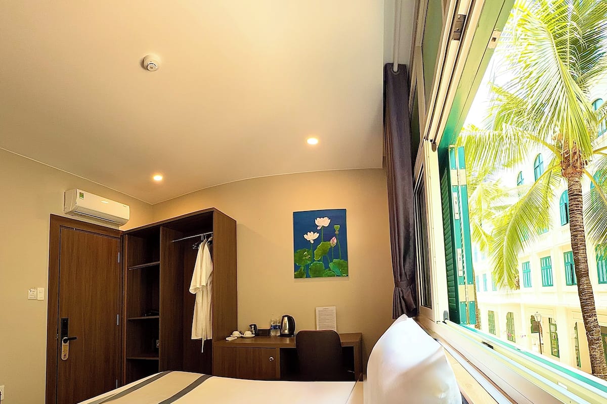 A hotel room with a bed, a desk, a wardrobe, a painting, and a window with a view of palm trees.