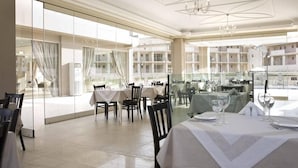 Un comedor con mesas dispuestas para dos, ventanas amplias y vista a un edificio.