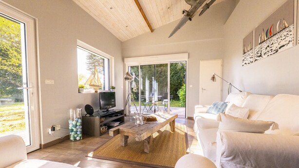 Ein modernes Wohnzimmer mit einer Couch, einem Couchtisch und einem Fernseher. Ein großes Fenster und eine Tür führen nach draußen.