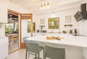 A modern kitchen with a large island, wooden chairs, a dining area with a table and chairs, and a view of greenery outside. - Quatre Vents (Sainte-Luce)