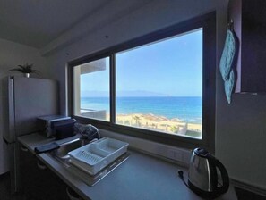 A kitchen with a large window overlooking a beach, a kettle, and a dish rack.