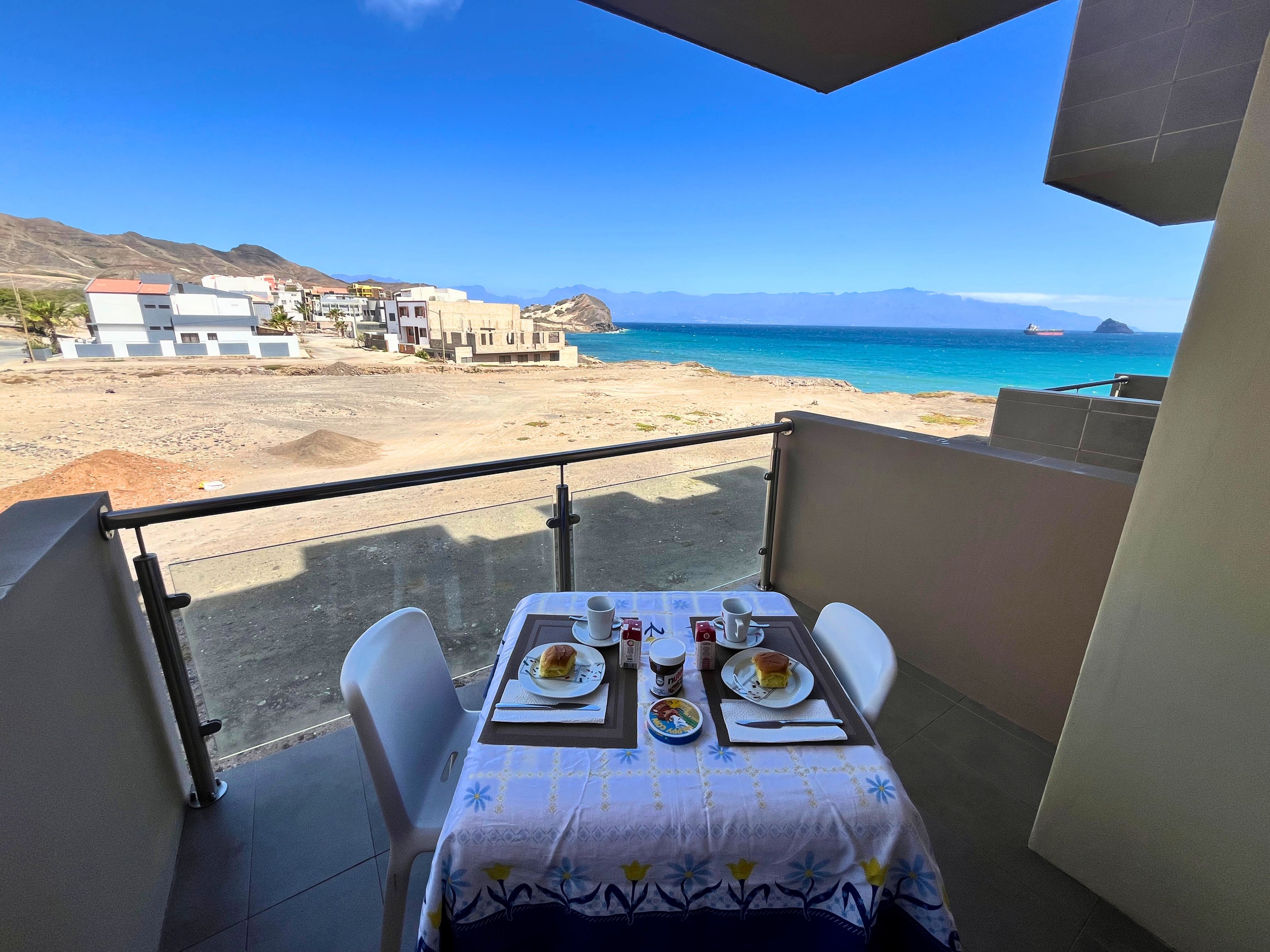 Un balcón con una mesa para dos, con vista a una playa y montañas.
