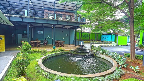 Un bâtiment doté d’un espace extérieur aménagé avec une zone de sièges couverte, d’une fontaine circulaire et d’une clôture verte en arrière-plan.