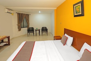 A hotel room with a bed, two chairs, a small table, an air conditioner, and a picture on the wall.