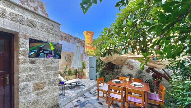 Ein Gartenbereich mit Steinwänden, einer gefliesten Terrasse, einem Holztisch mit Stühlen und einem Fernseher, der ein Rennspiel zeigt.