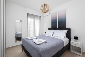 A modern bedroom with a bed, a bedside table, a hanging light fixture, and a large window with curtains. - Getaway Apartment in Rhodes Town (Rhodes)