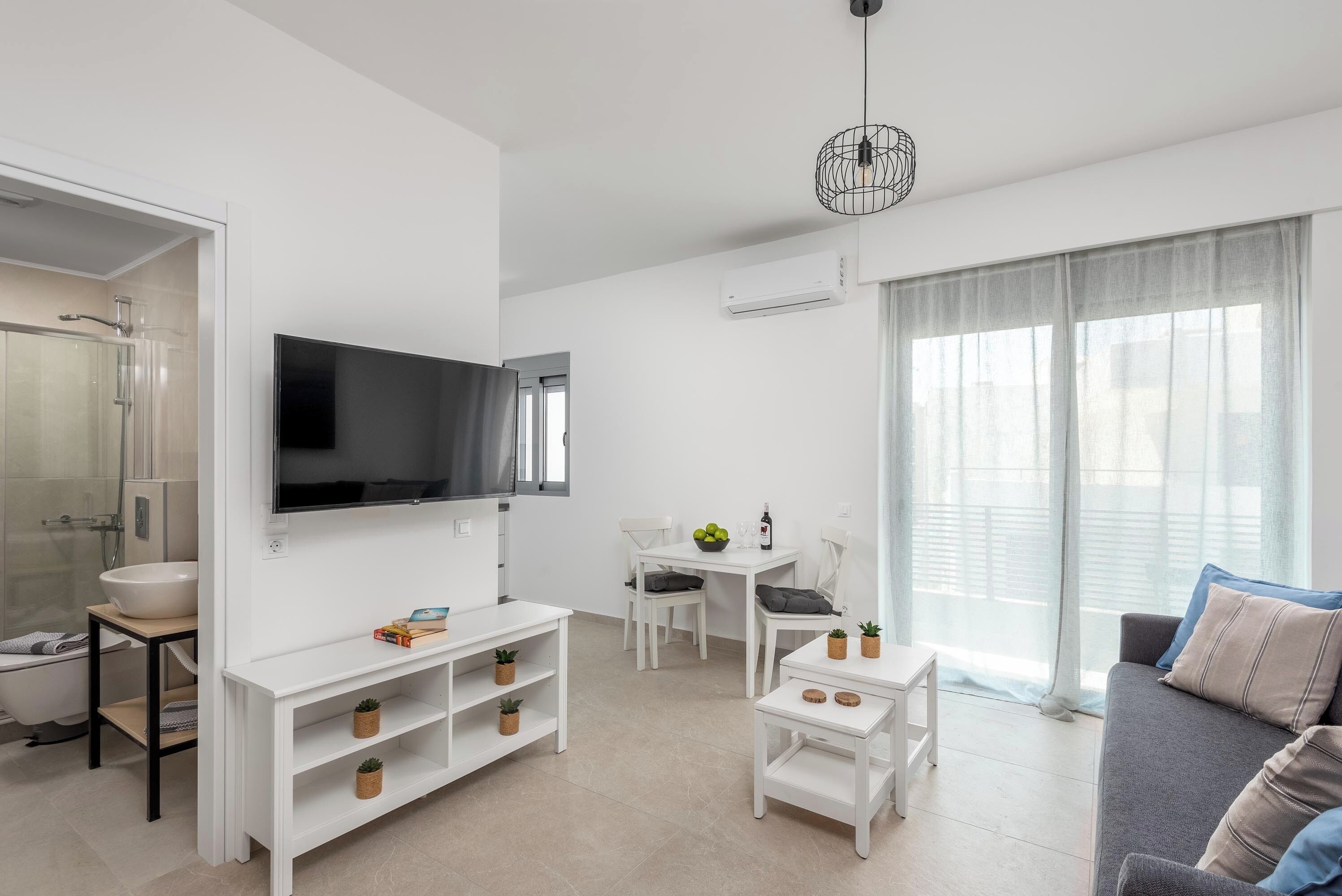 A modern living room with a flat-screen TV, white furniture, a small table with plants, and a sofa with cushions.