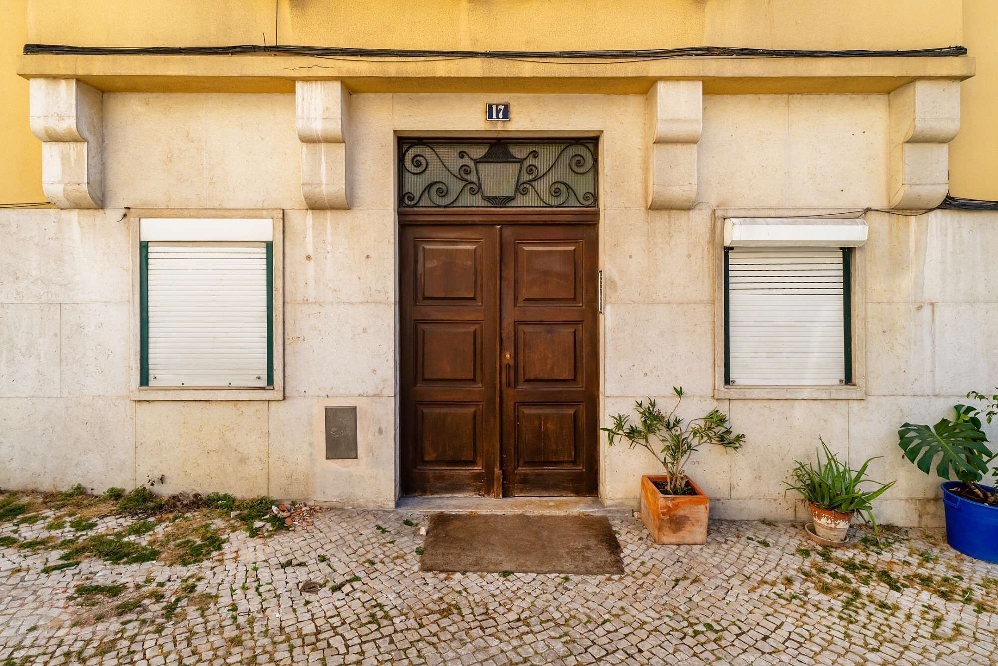 Una porta de fusta doble amb un marc decoratiu metàl·lic, flanquejada per dues finestres amb persianes tancades, i un paviment de pedra irregular al davant.