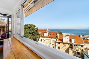 A room with a view of the sea, a wooden table, and a window with a view of buildings and a tree. - Sunny Terrace Apartment w/ River Views (Lisbon)