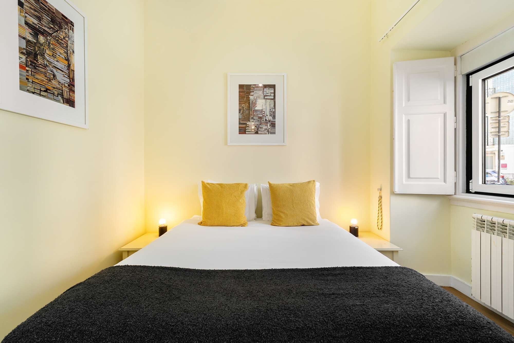 A hotel room with a bed, two bedside lamps, a framed picture on the wall, and a window with a view of buildings.