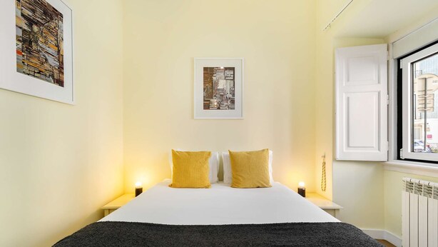 A hotel room with a bed, two bedside lamps, a framed picture on the wall, and a window with a view of buildings. - Sunny Terrace Apartment w/ River Views (Lisbon)