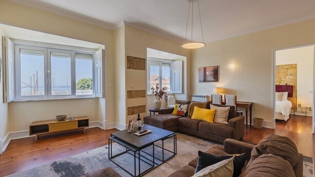 A living room with a sofa, coffee table, and a view of the sea through large windows. - Spacious Mordern Apt in Graça’s Historic Quarter (Lisbon)