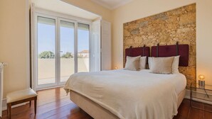 A bedroom with a large bed, a stone accent wall, a sliding glass door, and a bench. - Spacious Mordern Apt in Graça’s Historic Quarter (Lisbon)