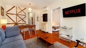 Een woonkamer met een bank, een houten salontafel, een aan de muur bevestigde televisie en een eethoek met een tafel en stoelen.