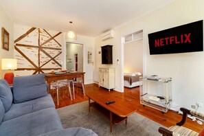 Ein Wohnzimmer mit einer Couch, einem Holztisch, einem an der Wand montierten Fernseher und einem Essbereich mit Tisch und Stühlen.