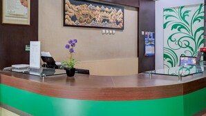 Un mostrador de recepción con una decoración en tonos verde y marrón, un cuadro enmarcado encima, una planta en maceta y un panel decorativo en la pared.