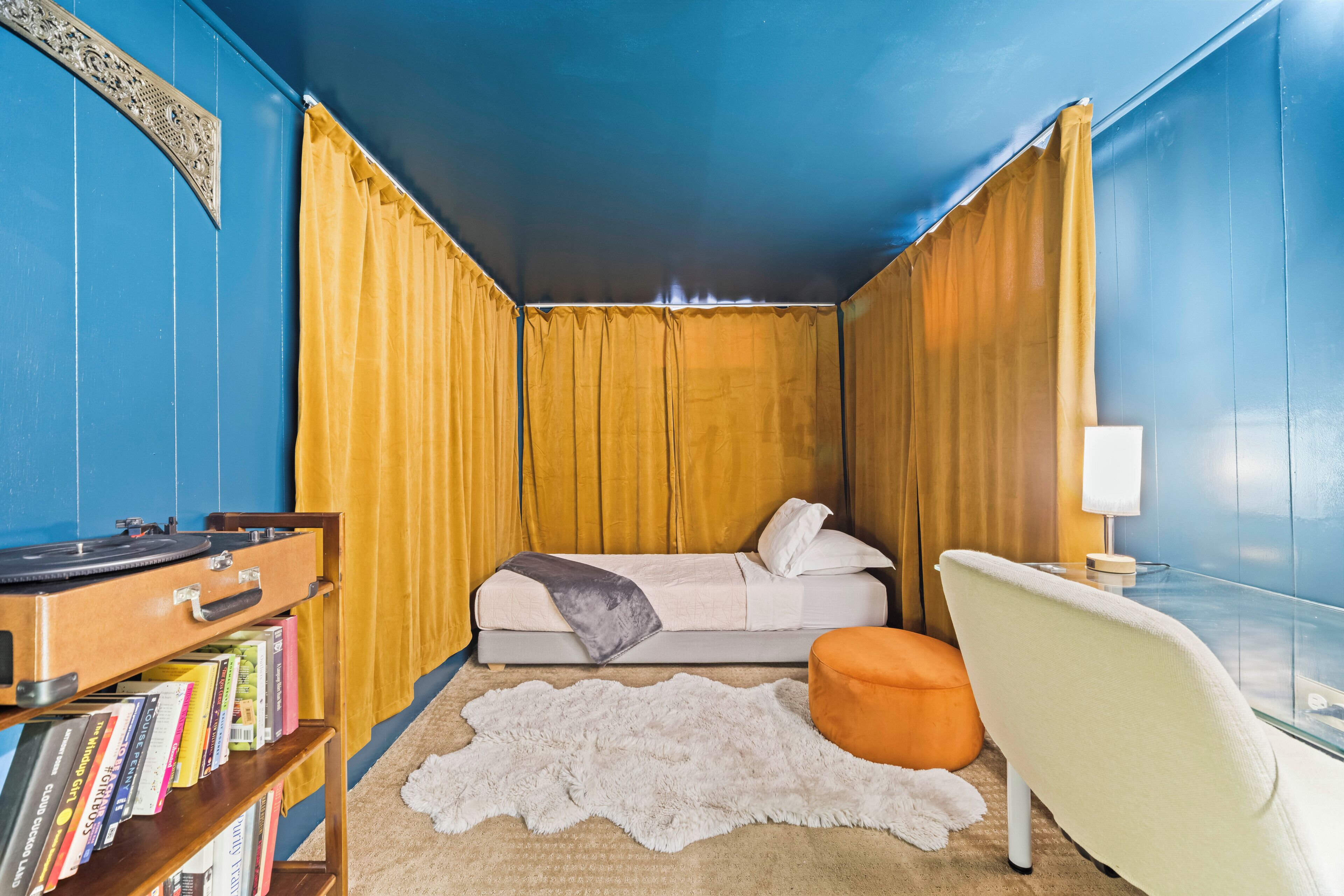 Ein Zimmer mit einem Bett, einem Sessel, einem kleinen Tisch, einer Lampe, einem Bücherregal und einem Teppich.