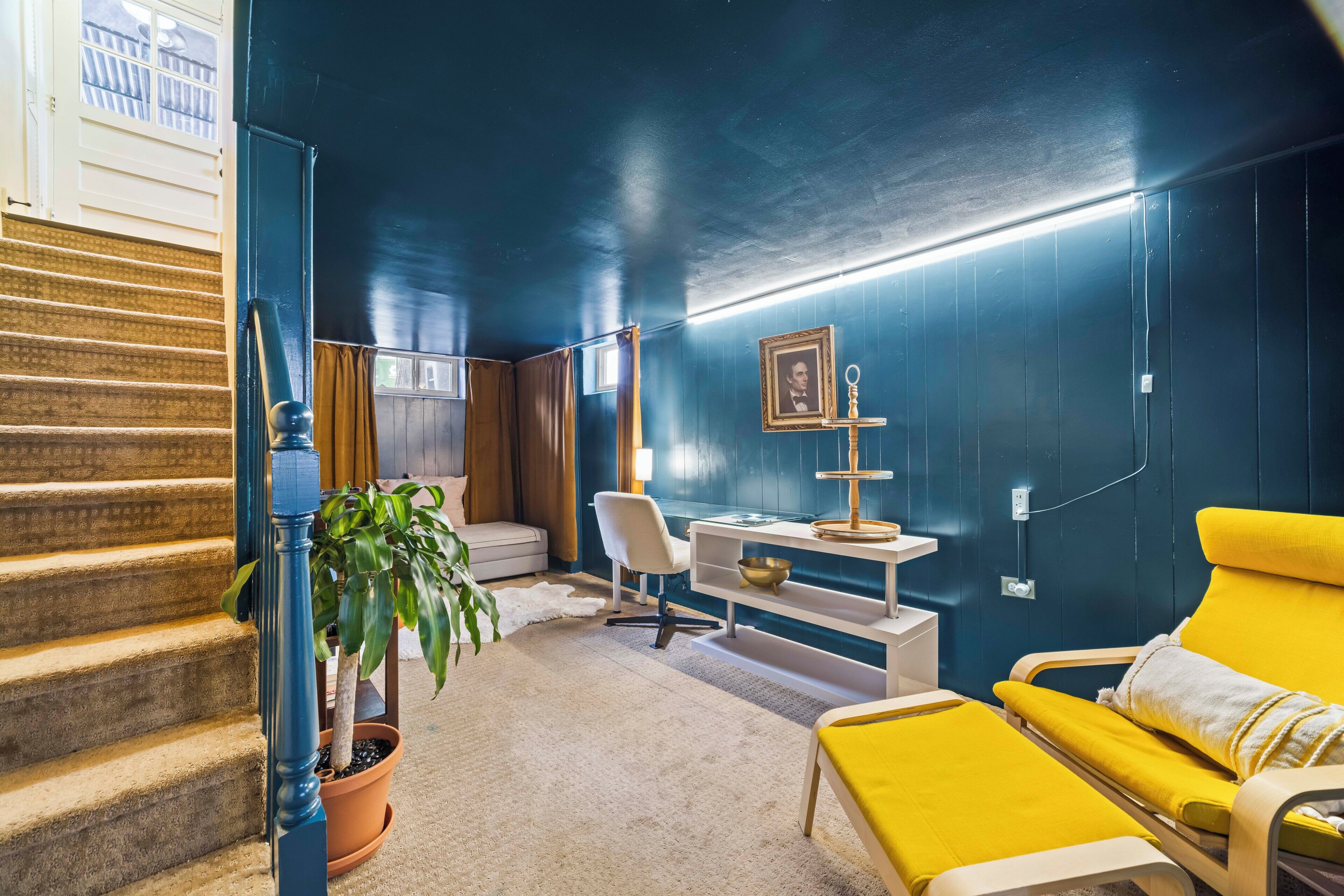 A room with a staircase, a desk, a yellow sofa, a potted plant, and a framed picture on the wall.