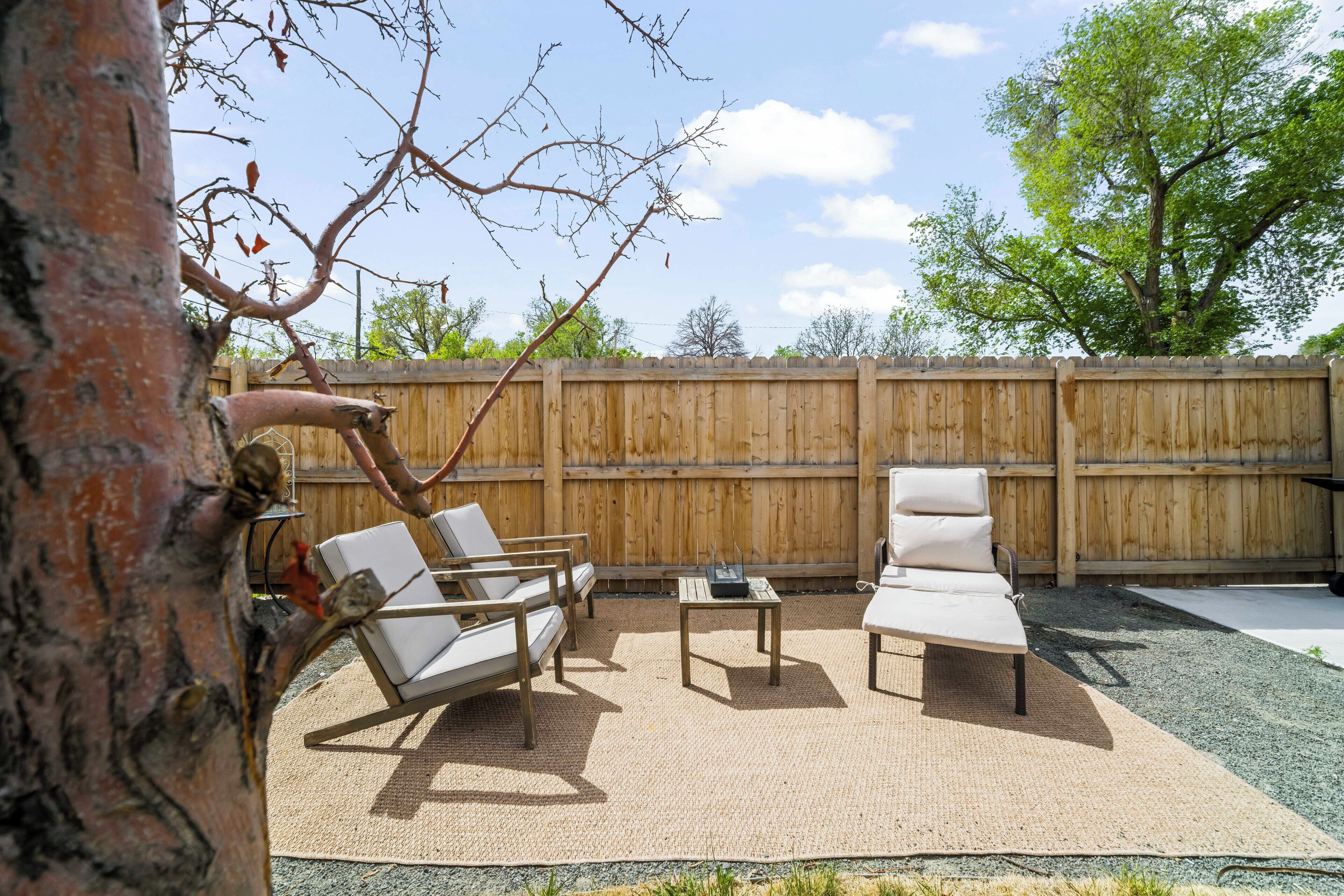 A backyard with a wooden fence, two white lounge chairs, a small table, and a tree.