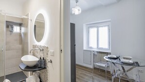 Un baño moderno con un espejo grande, un lavamanos y una ducha. Hay una zona de comedor con una mesa y sillas.