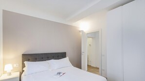 Una cama bien hecha con sábanas blancas, un cabecero tapizado en gris y una lámpara de noche.