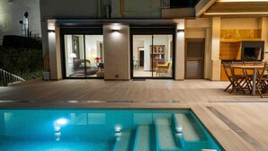 A swimming pool with a glass enclosure, a dining area with wooden furniture, and a kitchen area with a built-in stove.