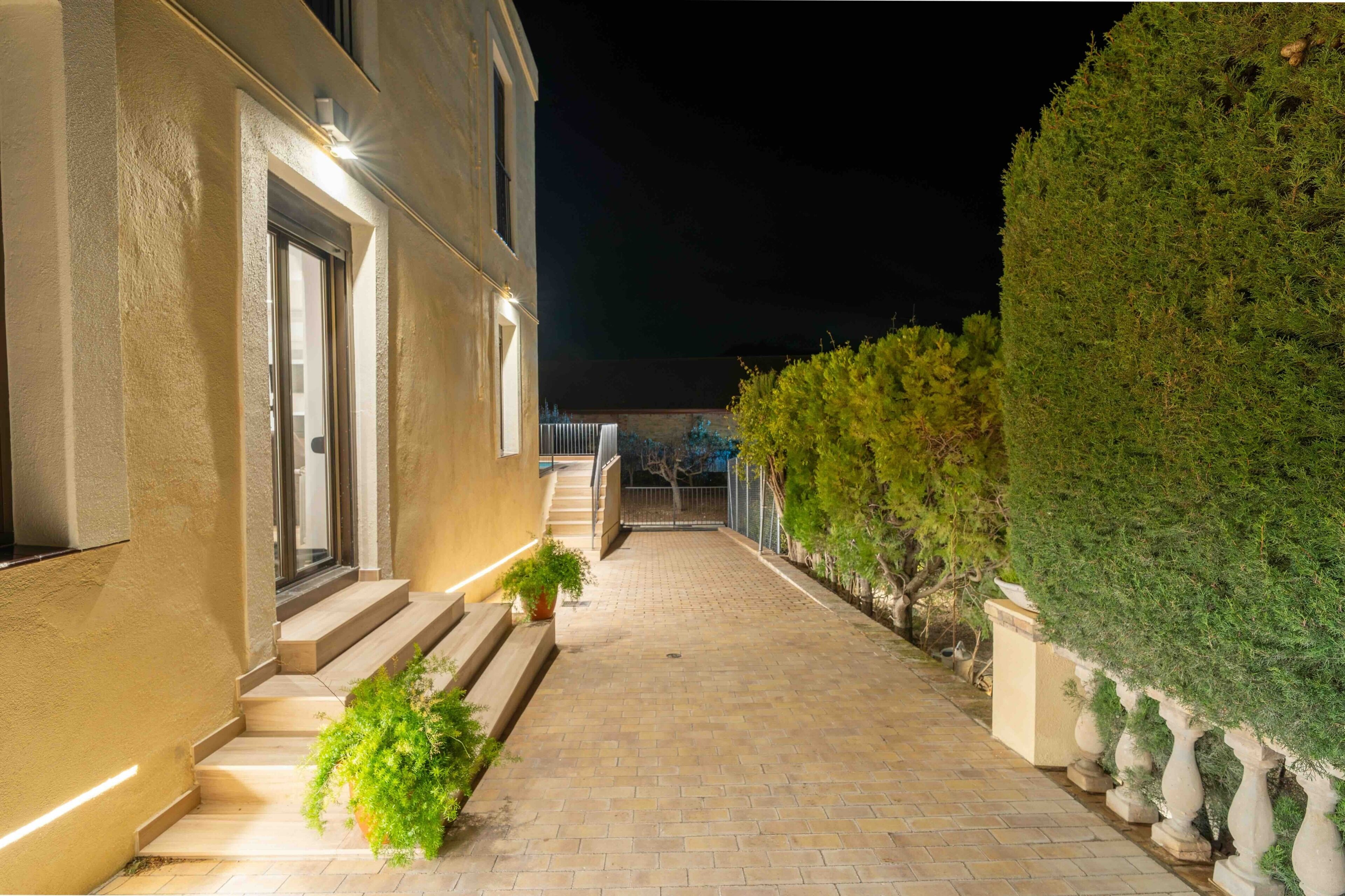Un sendero pavimentado que conduce a un edificio con escalones y una puerta, flanqueado por macetas con plantas y un seto bien cuidado.