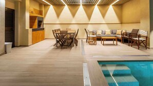 An indoor pool area with a wooden deck, a dining table with chairs, a sofa, and a kitchen area with cabinets.