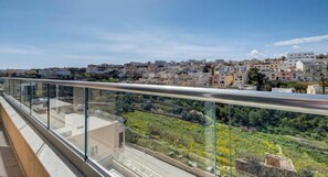 A balcony with a glass railing overlooking a cityscape with buildings and greenery. - Prime Swieqi 6BR w/ Private Pool & Sundeck (Swieqi)